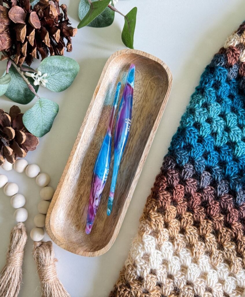 A wooden tray holds two colorful crochet hooks, surrounded by pinecones, eucalyptus leaves, a wooden bead garland, and a cozy crochet cardigan on a white surface.