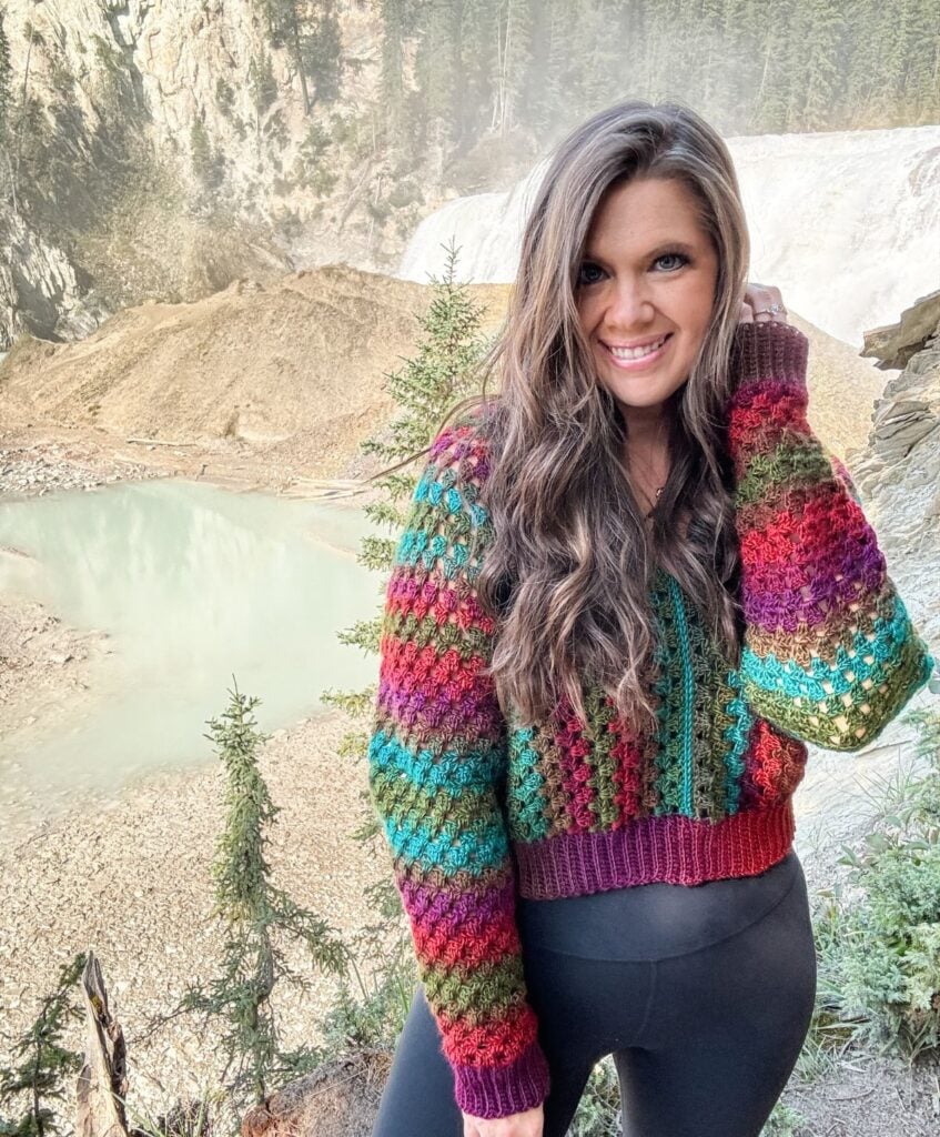 A woman is standing outdoors in front of a scenic landscape wearing a colorful crochet pullover. There is a body of water and trees behind her.