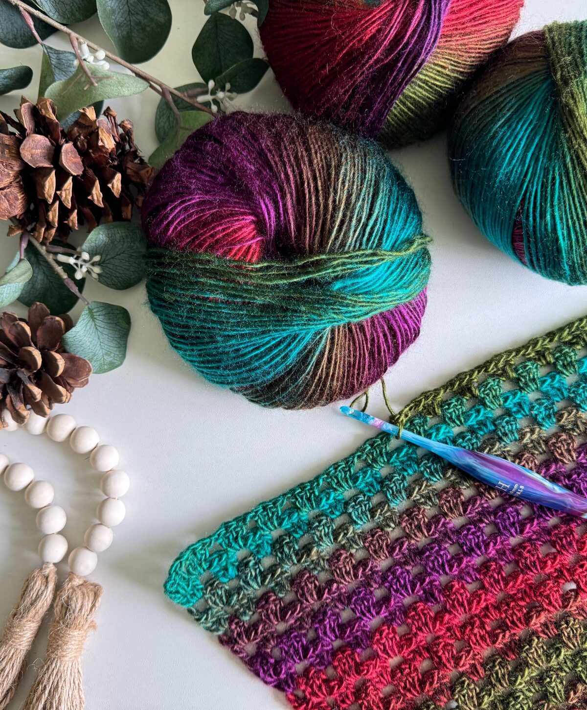 Yarn balls in multicolored hues with a partially crocheted piece, surrounded by pine cones, a crochet hook, and eucalyptus leaves on a white surface—a perfect setting for starting your next cozy crochet pullover.