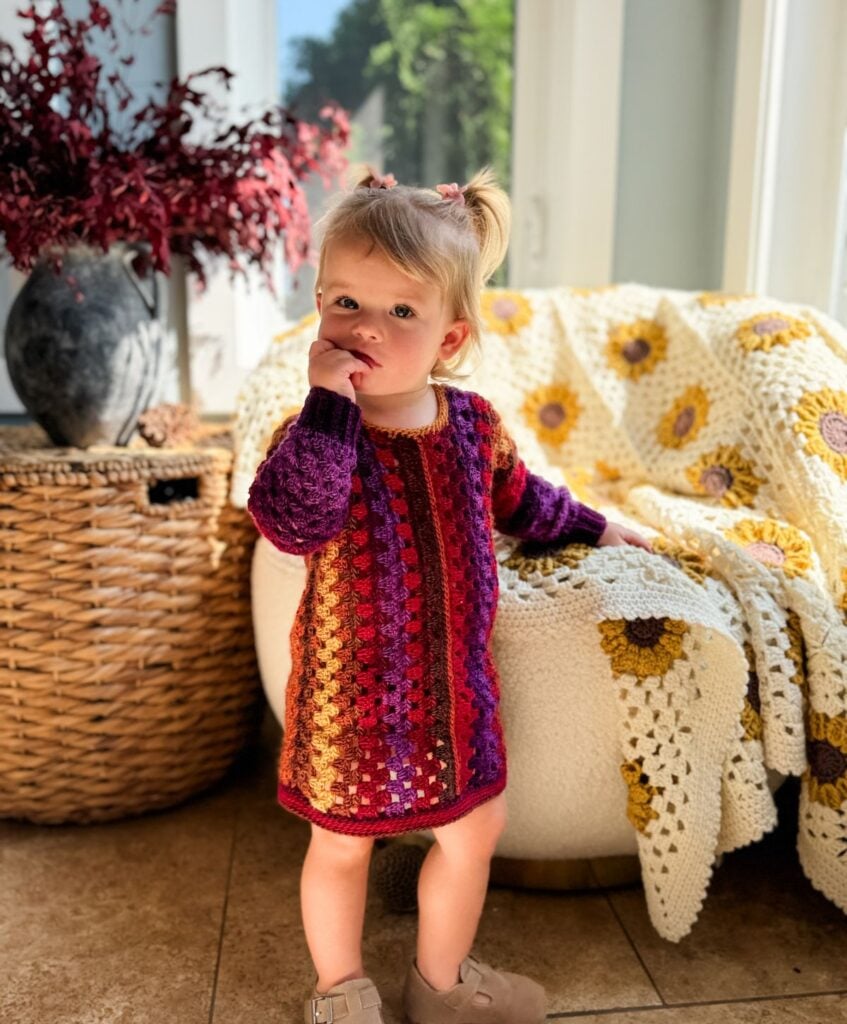 A toddler in a vibrant children's granny dress stands indoors, with a hand in her mouth. A patterned blanket and basket complete the charming scene.