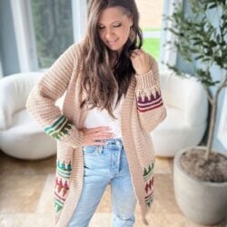 A woman in a mosaic crochet cardigan with colorful patterns, wearing a white top and ripped jeans, stands indoors near a plant and white chairs.