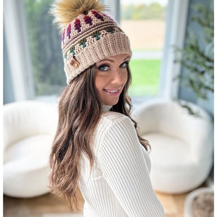 A woman in a white sweater smiles, wearing an Autumn's Charm crochet hat with a pom-pom. She stands in a bright room with white chairs and large windows.