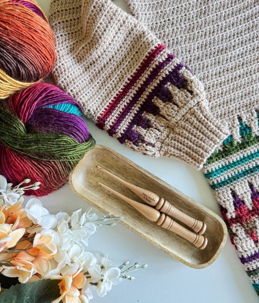 A mosaic crochet cardigan crafted from multicolored yarn lies beside crochet hooks in a wooden tray, accompanied by a bouquet of white and peach flowers.