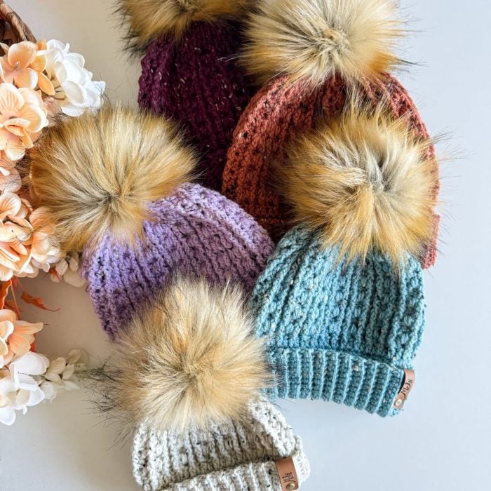 Five cozy crochet beanies with faux fur pom-poms in vibrant colors—purple, red, orange, blue, and beige—are artfully arranged on a surface next to a bouquet of light-colored flowers.
