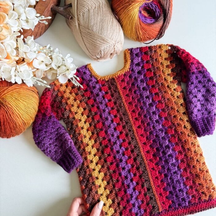 A hand holds a colorful crocheted sweater with a gradient pattern reminiscent of a children's granny dress, surrounded by yarn balls and white flowers.