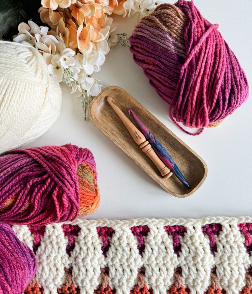 A wooden tray cradles crochet hooks, while colorful yarn balls and a partially completed mosaic crochet blanket lie beside it. In the background, flowers gently frame this vibrant crafting scene.