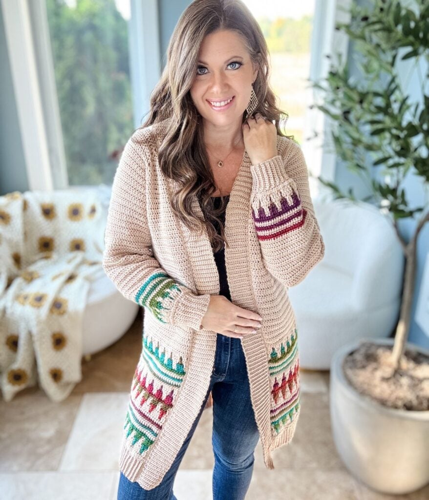A person stands indoors near a sofa and a potted plant, wrapped in a beige knitted cardigan with colorful patterns reminiscent of mosaic crochet work.