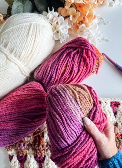 A hand holds pink and purple yarn balls next to white yarn and a mosaic crochet blanket in progress on a table adorned with floral decorations.