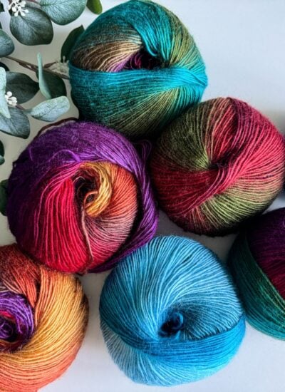 Six balls of multicolored yarn displayed on a white surface with a sprig of green leaves on the side, perfect for crafting a vibrant mosaic crochet cardigan.