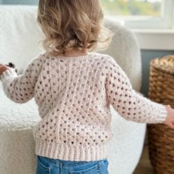 A toddler with curly hair wears a knitted sweater reminiscent of a children's granny dress and jeans, standing in a room with a soft chair and a wicker basket.