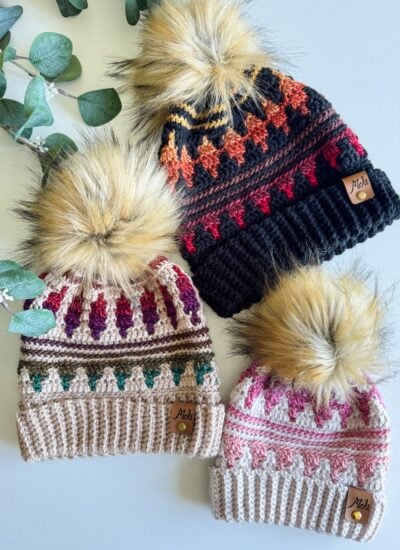Three knitted hats, including the Mosaic Crochet Hat with pom-poms and colorful patterns, are artfully arranged on a white surface next to green eucalyptus leaves.