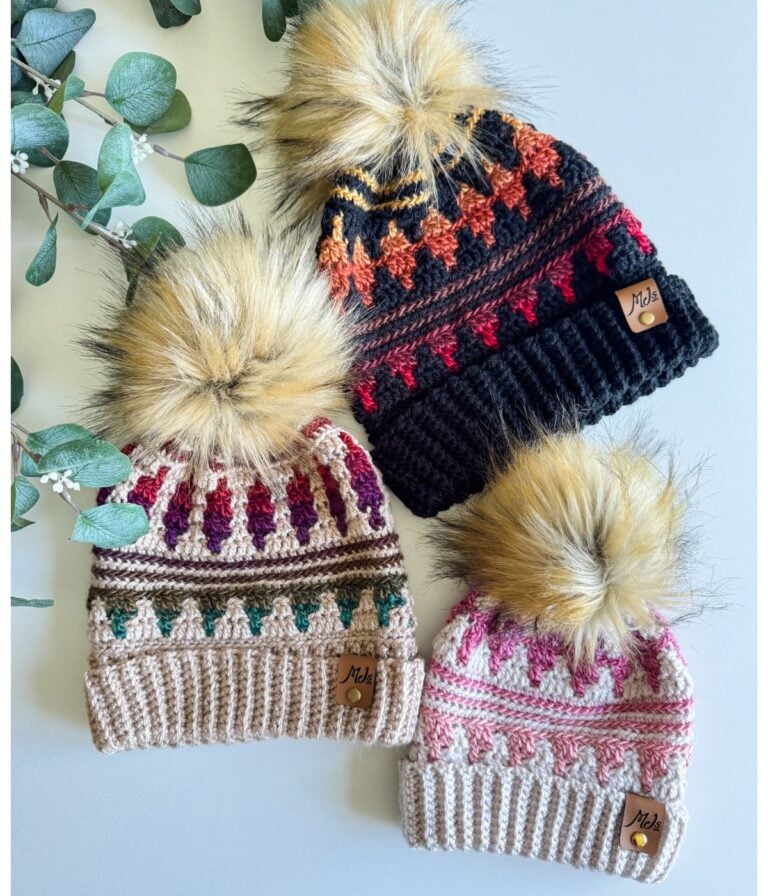 Three knitted hats, including the Mosaic Crochet Hat with pom-poms and colorful patterns, are artfully arranged on a white surface next to green eucalyptus leaves.