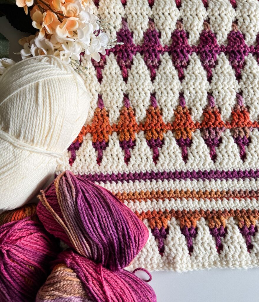Mosaic crochet blanket featuring a geometric pattern in white, purple, and orange tones. Paired with skeins of white and multicolored yarn, complemented by vibrant orange flowers.