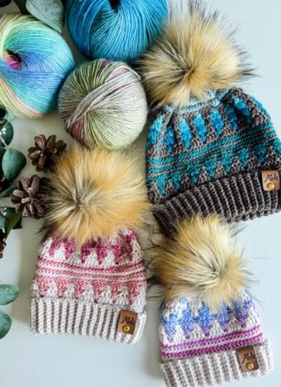 Three knitted hats with pom-poms, in pink, blue, and green patterns, embody Autumn's Charm. They are surrounded by balls of yarn, pinecones, and eucalyptus leaves on a light surface.