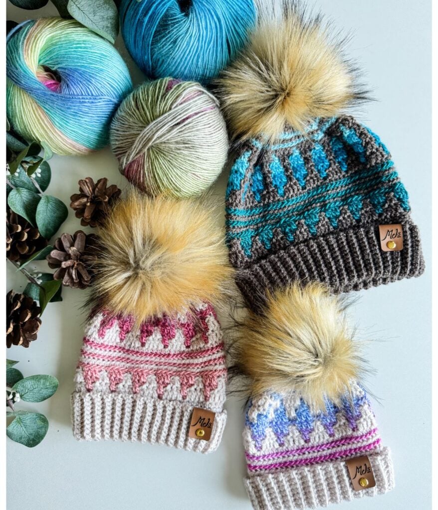 Three knitted hats with pom-poms, in pink, blue, and green patterns, embody Autumn's Charm. They are surrounded by balls of yarn, pinecones, and eucalyptus leaves on a light surface.