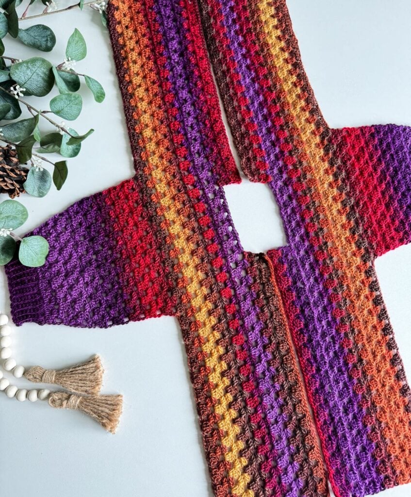 A vibrant children's granny dress, crocheted in shades of red, orange, purple, and brown, lies flat with a rectangular opening. On the side sit green leaves and a playful decorative tassel.