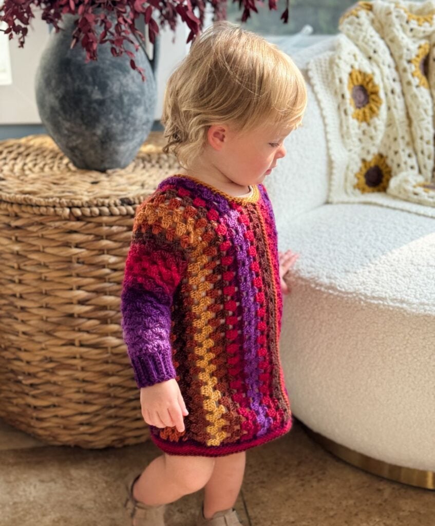 A toddler wearing a vibrant children's granny dress stands next to a woven basket and a white textured chair.