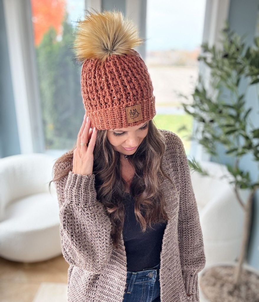 A woman wearing a brown crochet beanie with a pom-pom and a matching cardigan stands indoors near a window.