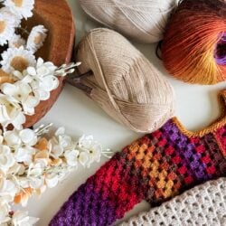 A crocheted sweater and a fabric piece, alongside colorful yarn balls, adorn the table. Nearby, a wooden bowl brims with delicate white flowers, creating a whimsical scene that evokes the charm of a children's granny dress.