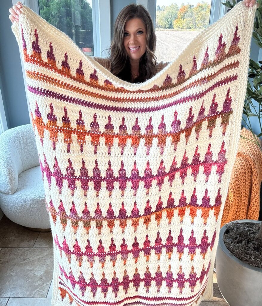 A person drapes a crocheted blanket with a geometric pattern in shades of purple, orange, and white, matching their cozy crochet pullover.