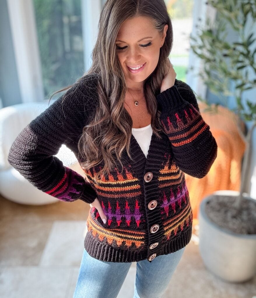 A person wearing a mosaic crochet cardigan with colorful patterns and jeans stands indoors near a plant.