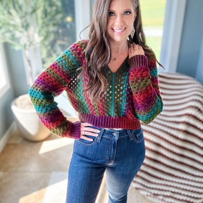 A woman with long hair smiles, wearing a colorful crochet pullover and jeans, standing in a well-lit room with large windows and a green plant in the background.