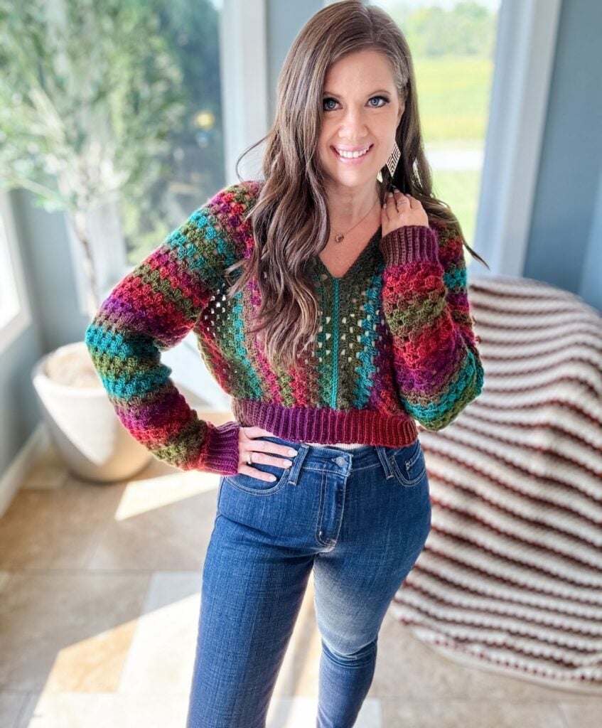 A woman with long hair smiles, wearing a colorful crochet pullover and jeans, standing in a well-lit room with large windows and a green plant in the background.