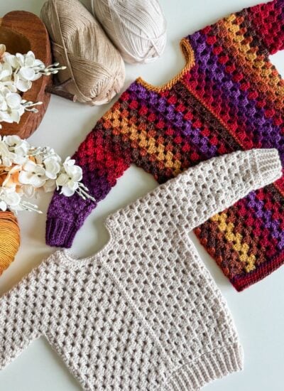 Two crocheted sweaters, including a vibrant children's granny dress in shades of red, orange, purple, and yellow, are displayed on a table alongside a beige sweater. Nearby lie balls of yarn and decorative flowers.