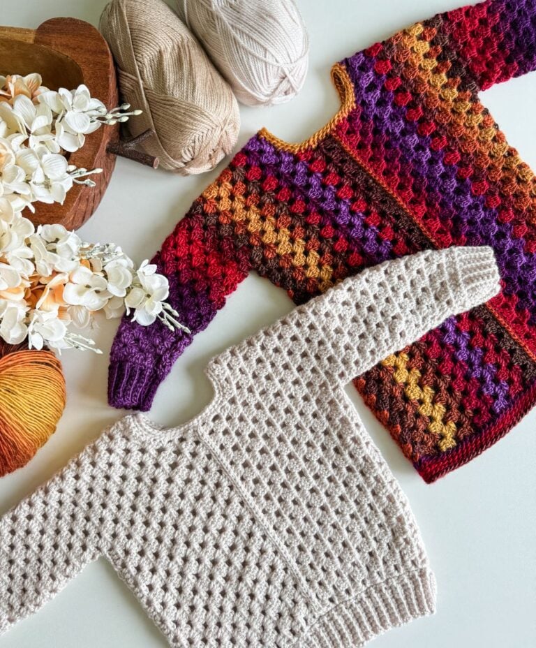 Two crocheted sweaters, including a vibrant children's granny dress in shades of red, orange, purple, and yellow, are displayed on a table alongside a beige sweater. Nearby lie balls of yarn and decorative flowers.