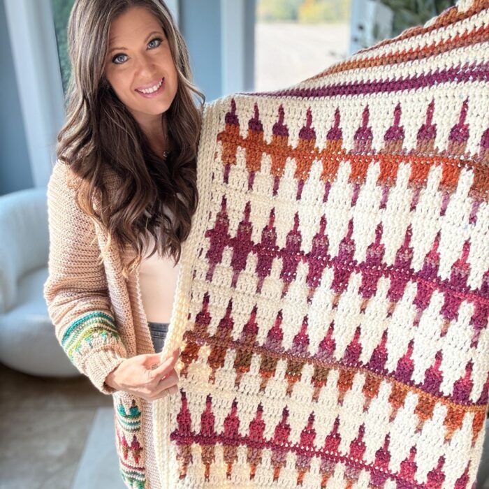 Person holding a vibrant creation, a crocheted blanket with a geometric pattern of triangles in red, orange, and purple hues. The craftsmanship rivals that of a finely woven crochet pullover, blending creativity and texture into an exquisite textile masterpiece.
