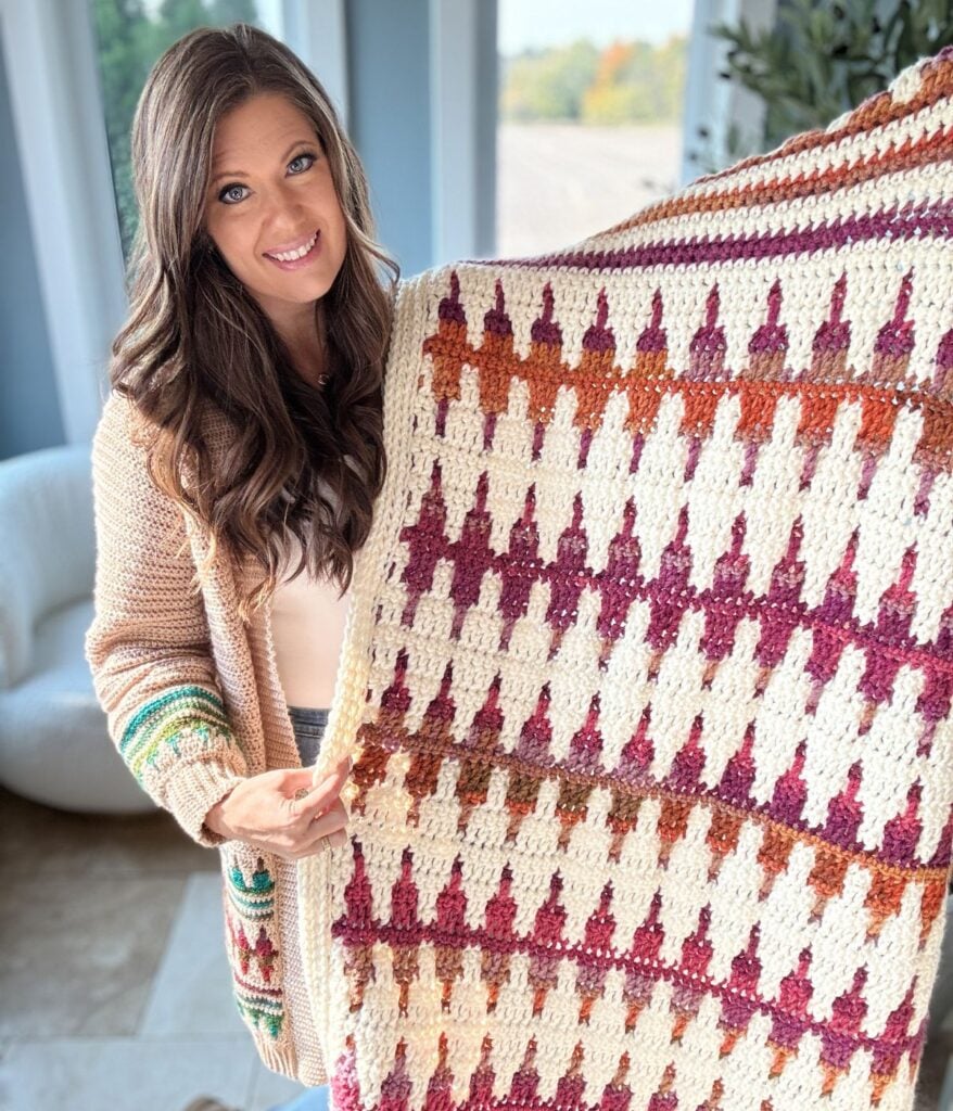 Person holding a vibrant creation, a crocheted blanket with a geometric pattern of triangles in red, orange, and purple hues. The craftsmanship rivals that of a finely woven crochet pullover, blending creativity and texture into an exquisite textile masterpiece.