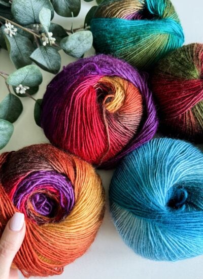 Five skeins of colorful yarn in shades of red, purple, blue, and green sit near eucalyptus leaves. A hand gently touches one skein, perhaps envisioning them transformed into a playful children's granny dress.