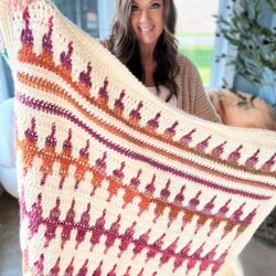 A woman holds a colorful, patterned crochet blanket inside a room with large windows, where the light softly highlights her matching crochet pullover.