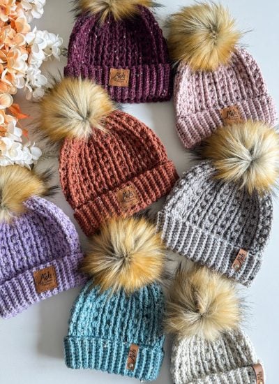 Seven cozy crochet beanies with faux fur pom-poms in a variety of colors are arranged on a surface next to a cluster of orange and white flowers.