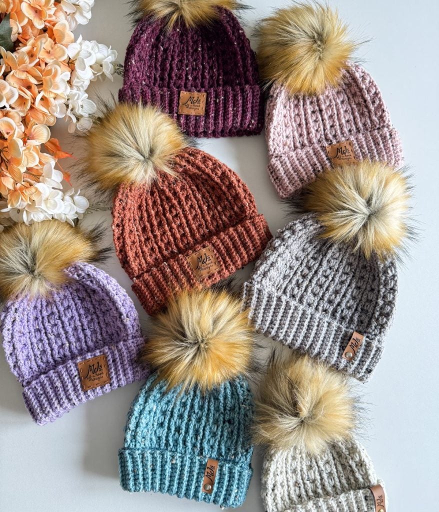 Seven cozy crochet beanies with faux fur pom-poms in a variety of colors are arranged on a surface next to a cluster of orange and white flowers.