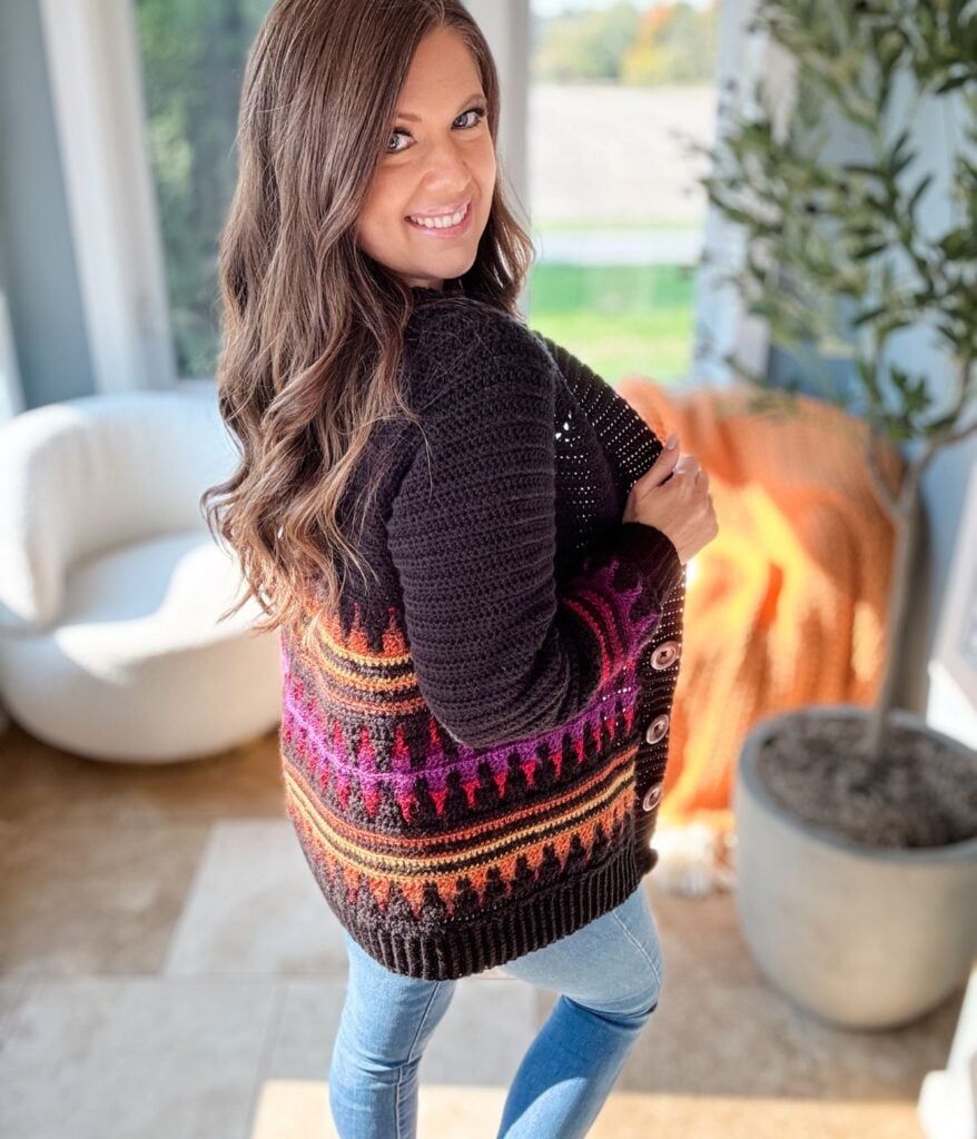 A person with long hair smiles while wearing a colorful mosaic crochet cardigan and jeans, standing in a sunny room with a white chair and an orange blanket in the background.