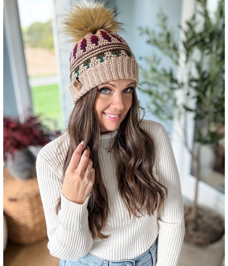 A woman wearing a knitted crochet hat with a pom-pom, cream sweater, and jeans, smiles indoors with plants in the background, capturing Autumn's Charm effortlessly.