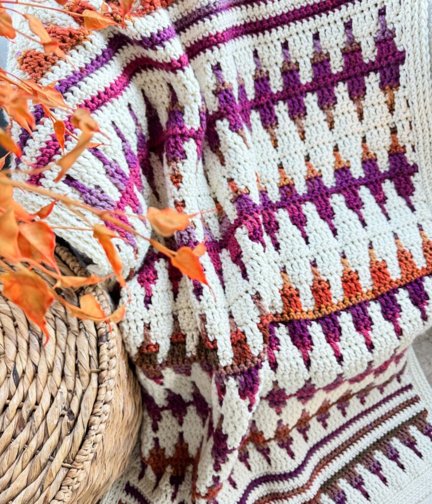 A mosaic crochet blanket with geometric patterns in purple, orange, and white is draped over a woven basket. Orange leaves are visible at the top left.
