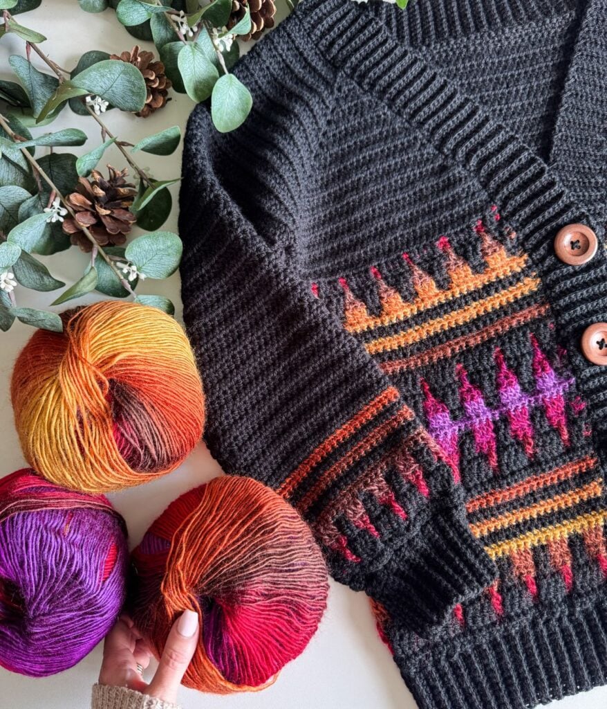 A colorful knitted sweater lies next to three balls of gradient yarn in orange, purple, and red tones, while a hand touches one yarn ball. The scene resembles a mosaic crochet cardigan with greenery and pinecones in the background.