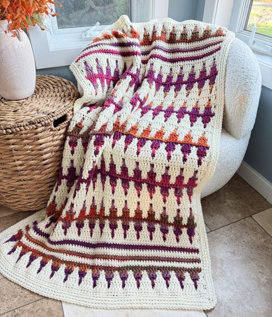 A vibrant mosaic crochet blanket with geometric patterns is draped over a light-colored chair, perfectly complementing the nearby woven basket.