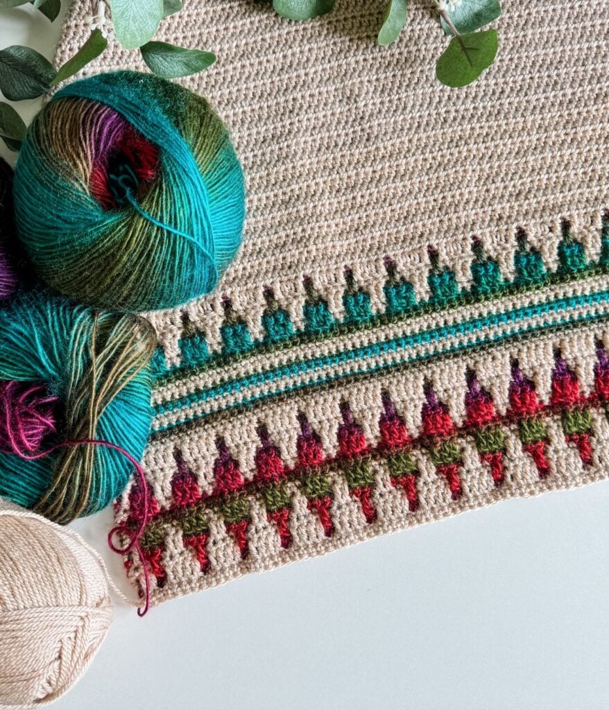 A mosaic crochet cardigan features vibrant geometric patterns, enhanced by balls of yarn in green, blue, and purple hues. Eucalyptus leaves add a touch of nature in the corner.