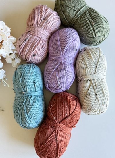 Six skeins of yarn in pink, green, purple, beige, blue, and rust orange are arranged on a surface next to a cluster of white and peach flowers—perfect for crafting a vibrant crochet beanie.