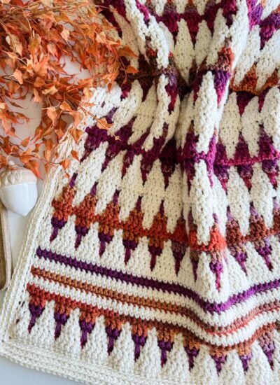 A mosaic crochet blanket with geometric patterns in shades of orange, red, and purple lies next to autumn leaves, a wooden spindle, and an acorn decoration.