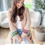 A woman wearing a beige cardigan with vibrant mosaic crochet patterns stands in a bright room with a white armchair and potted plant. Text on the image promotes this stylish top-down crochet cardigan pattern.