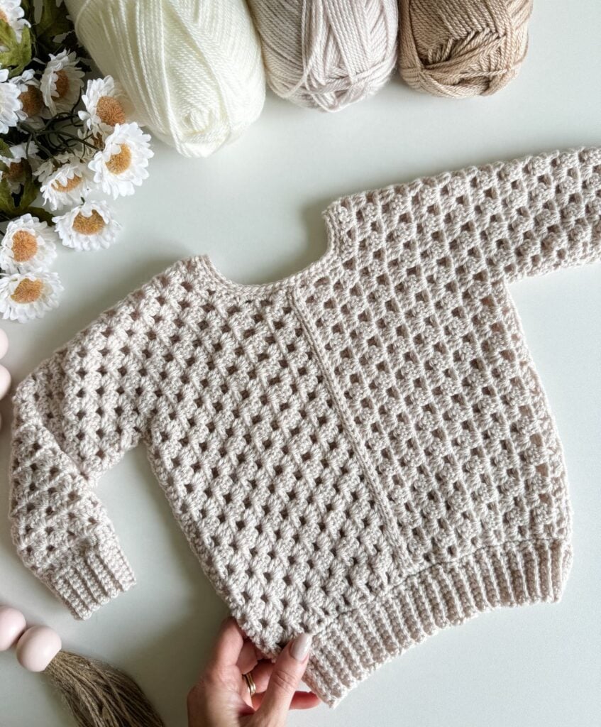 A hand holds a knitted beige baby sweater, reminiscent of a children's granny dress, surrounded by white and brown yarn and delicate white flowers on a light-colored surface.