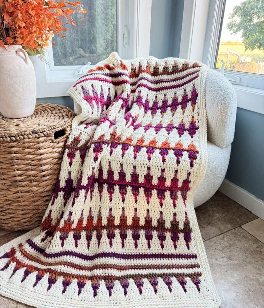 A Mosaic Crochet Blanket with a geometric pattern in cream, purple, and orange is draped over a white chair beside a woven basket and window.