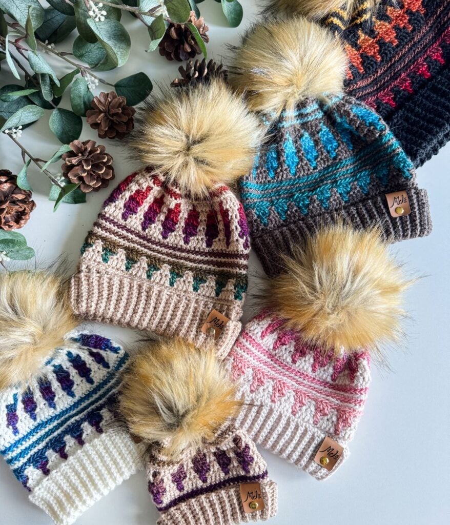 Several mosaic hats with colorful patterns and large faux fur pom-poms are arranged on a table next to pinecones and green leaves.