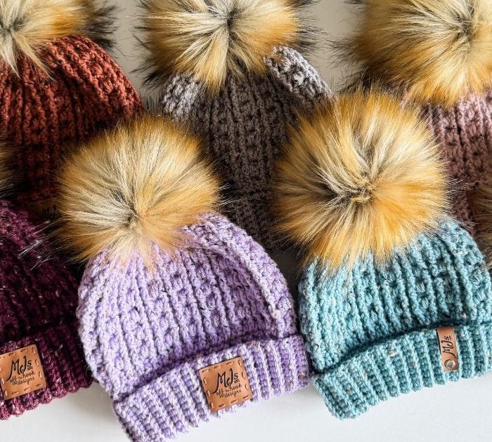 A row of seven vibrant crochet beanies, each adorned with a large faux fur pom-pom, displayed elegantly on a pristine white surface.