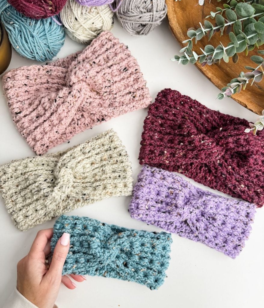 Four crochet headbands in pink, red, cream, and purple are arranged on a table with a hand holding a blue one. Yarn balls and green foliage form the background.