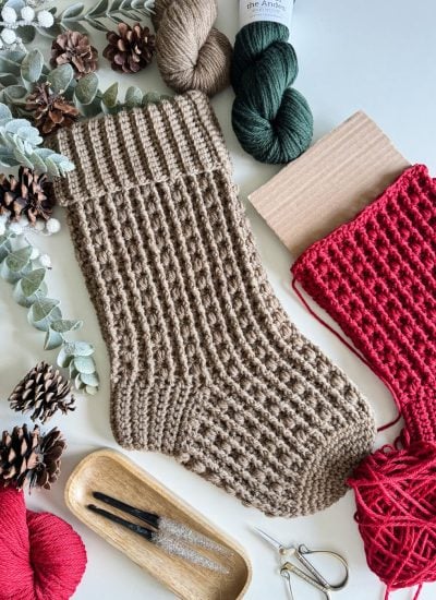 A knitted brown sock, a partially knitted red sock, yarn balls, knitting needles, and decorative foliage are arranged on a white surface, evoking the cozy ambiance of Snuggle Season Stocking projects.